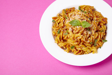 A dinner dish full of tomato and basil fusilli pasta on a bright pink background with blank space at side
