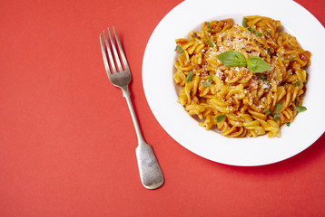 A dinner dish full of tomato and basil fusilli pasta on a bright red background with fork and blank space at side