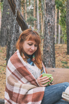 Beautiful red haired young woman with blanket and cup of coffee on a bench at the autumn park. Coloring and processing photo