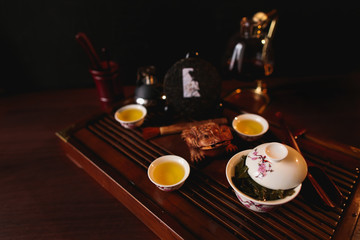 The Chinese tea items. Chinese tea set. Porcelain gaiwan, three cups with tea, pancake of traditional Chinese puer tea, siphon and golden frog on tea desk. Soft selective focus.