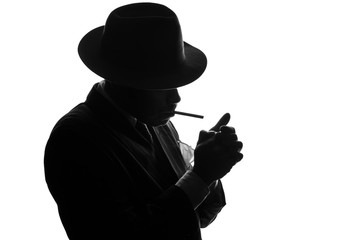 Silhouette of private detective lights cigarette. Agent looks like Al Capone stay side to camera. Police criminal scene in black and white. Gangster studio shot