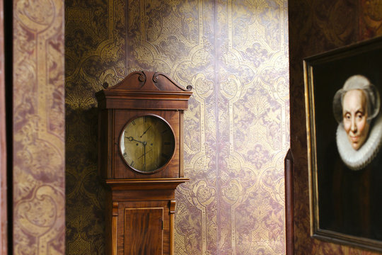 A classical clock in a corner againt a red and golden wallpaper and a painting of a woman.