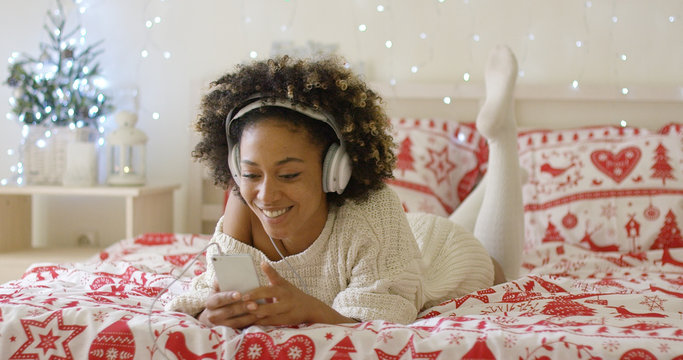 Attractive young woman relaxing at Christmas lying on her bed listening to music on her mobile with sparkling festive lights in the background