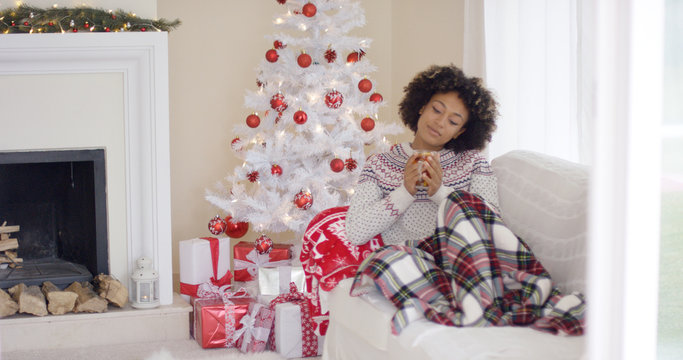 Young woman relaxing during the Christmas holiday season sitting under a rug on a sofa in front of the decorated tree with a cup of hot beverage.