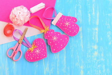 Christmas tree ornaments. Homemade pink felt Christmas tree, heart, mitten decorated with beads and snowflakes. Felt, scissors, pincushion, thread, needle on blue background with copy space for text