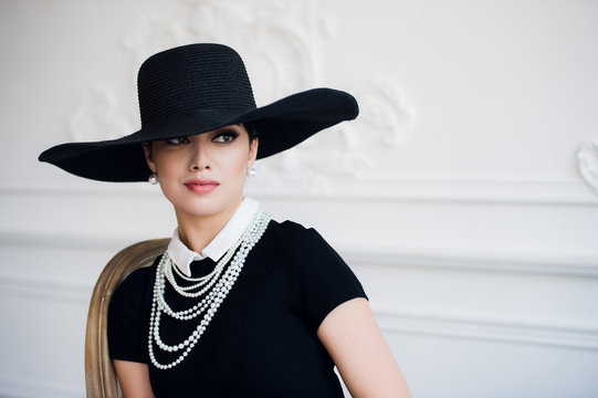 Portrait of a beautiful young woman in retro style in an elegant black hat and dress over luxury rococco wall background