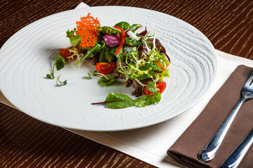 Vegetable salad menu for the restaurant in a large white plate on a white table cloth on a wooden table. Salad on the table with knife and fork. Svezhive vegetables.
