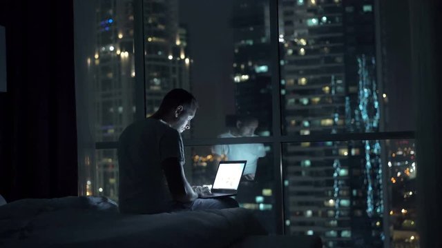 Silhouette of man using laptop and falling asleep on bed at night at home, 4K
