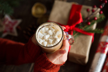 Closeup on hot chocolate with marshmallows in hand of happy woman against the background of a table with Christmas gifts

