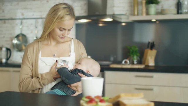 Beautiful Young Mother Feed Her Cute Child from a Bottle. All this is Happening on a Modern Kitchen. Shot on RED Cinema Camera in 4K (UHD).
