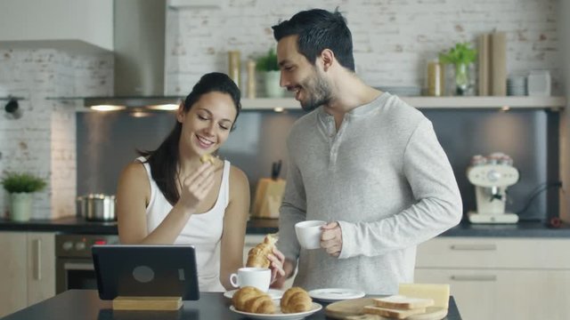Handsome Couple in the Kitchen with Tablet on the Table. She eats Croissant He Drinks Coffee.  Shot on RED Cinema Camera in 4K (UHD).