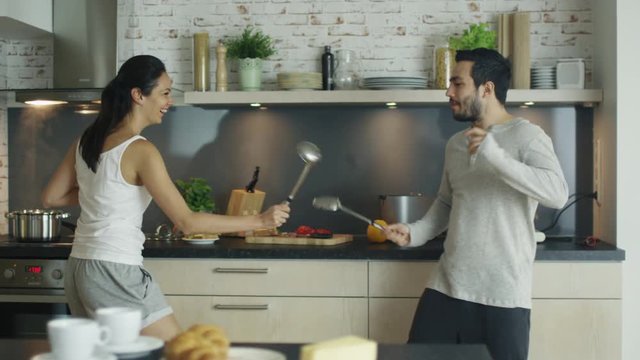 On the Kitchen Young Handsome Couple is Fencing with Kitchen Appliances. On the Stove Something Cooks in the Pan. Shot on RED Cinema Camera in 4K (UHD).