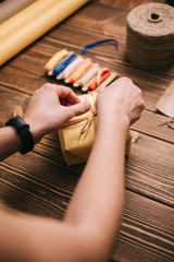 From above shot of crop hands decorating wrapped present with ribbon on wooden desk.