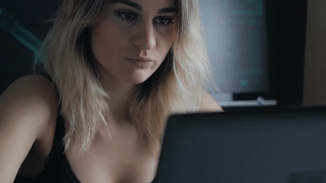 Blonde woman hacker working at a laptop with cup of coffee in dark room