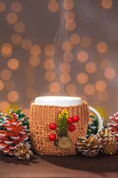 Closeup of white steaming winter cup in brown knitted sweater with christmas decorations on wooden table.