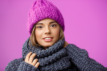 Young beautiful fair-haired girl in knited hat and sweater smiling looking at camera over violet background. 