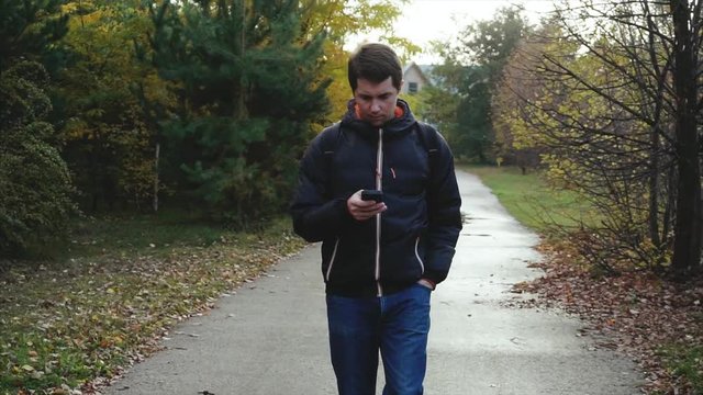 Young man in black jacket walking in the autumn park using his smartphone HD