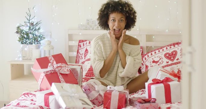 Excited young woman sitting on a bed full of Xmas gifts on a festive counterpane as she celebrates Christmas at home