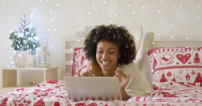 Gorgeous  happy and single young adult woman laying down in bed with computer while surrounded by Christmas tree lights. She video chatting with her friends or family.