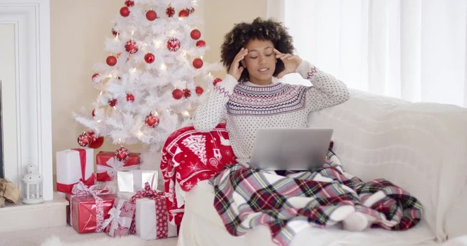 Young woman relaxing in front of the Christmas tree sitting under a rug on the sofa using her laptop computer gesturing with her hands