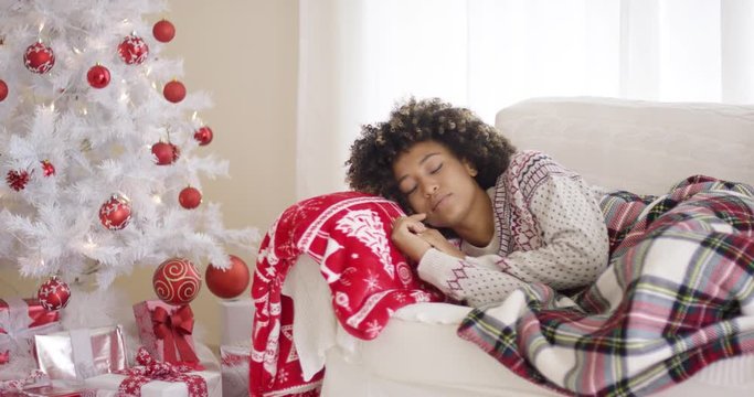 Single Black woman in sweater and plaid blanket sleeping on couch beside white Christmas tree