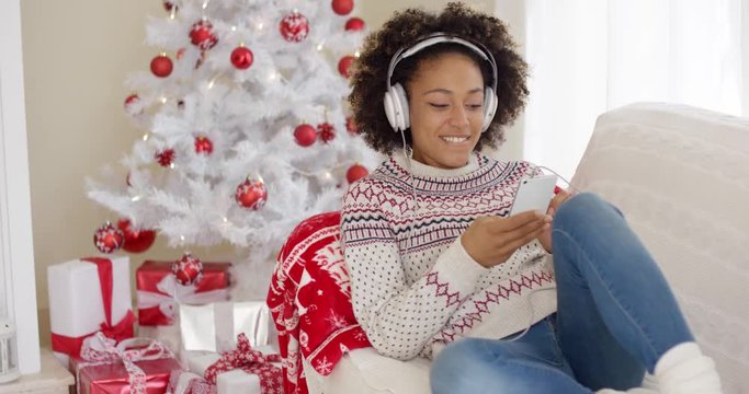 Attractive woman listening to music on her mobile phone at Christmas as she relaxes on a sofa in front of the tree with a happy smile.