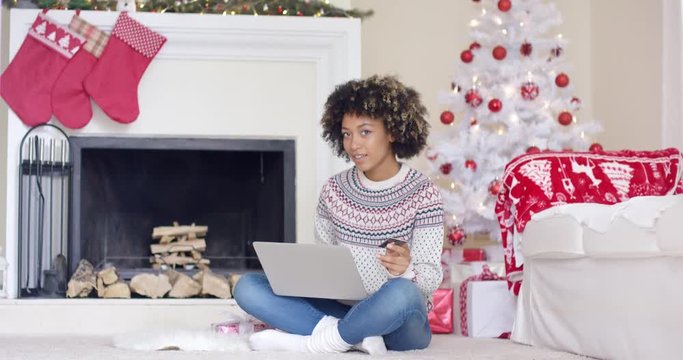 Young woman doing Christmas shopping online sitting on the floor in front of the tree with her laptop and credit card looking at merchandise on offer with a thoughtful expression.
