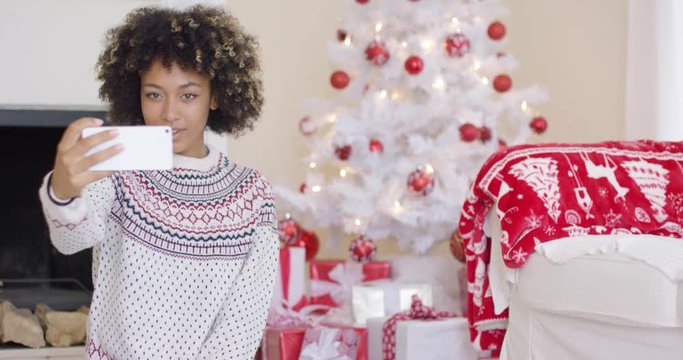 Happy young woman posing for a Christmas selfie on her mobile phone in front of a red and wite themed tree and gifts in a festive living room.