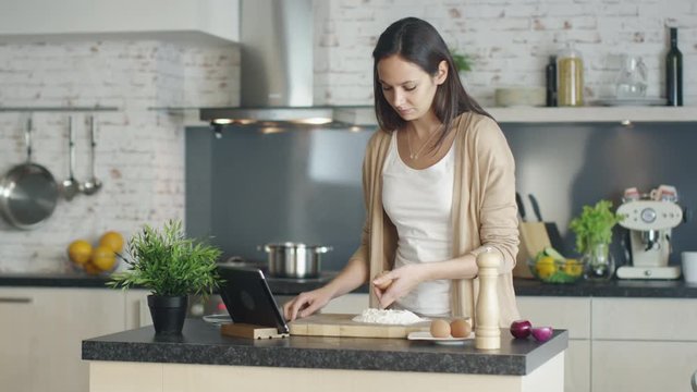 Young Girl is Cooking on the Kitchen. She for Recipe Reference on Her Tablet Computer and Breaks an Egg into a Heap of Measured Flour. Shot on RED Cinema Camera in 4K (UHD).