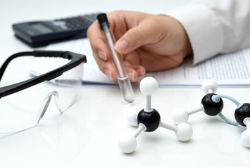 A scientist hand holding a test tube with liquid inside in the science laboratory with methane and ethylene molecular structure, safety glasses, calculator and experimental report on the table