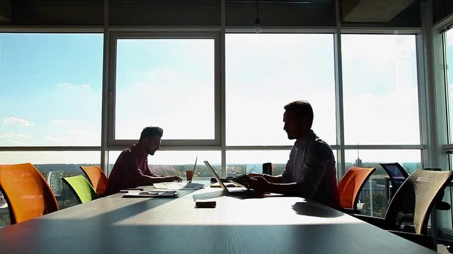 Businessman reading book during work ti,e while his colleague working on laptop computer in board room. Men communicating, smiling. Business concept.