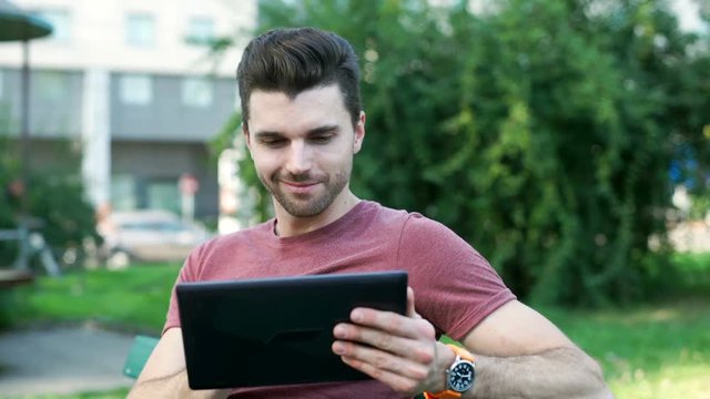 Handsome man sitting in the park and browsing internet on tablet
