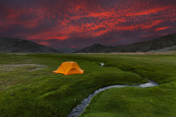 Orange tent at a small creek on the background of mountains and