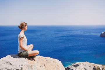 Fototapeta na wymiar Young woman relaxing outdoors. Attractive girl sitting on the rock and looking at sea. Ralaxation and active lifestyle concept