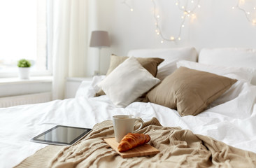 tablet pc, coffee cup and croissant on bed at home