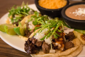 Plate of three street style tacos topped with micro greens, onions, chives and various sides