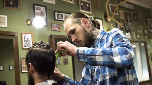 Attractive young barber is cutting human hair with the scissors. He is looking at hair with concentration. The bearded man is raising his chin