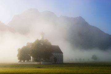 Fototapeta na wymiar Saint Coloman church with warm light shining through the fog during amazing sunrise near Neuschwanstein Castle, Fussen, Bavaria, Germany