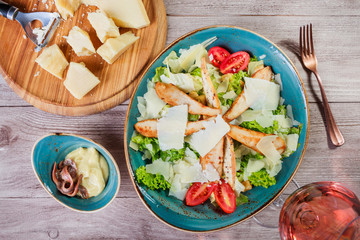 Salad with chicken breast, parmesan cheese, croutons, tomatoes, mixed greens, lettuce and glass of wine on light wooden background. Ingredients on table