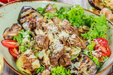 Hot salad with veal, mushrooms, salad leaves, eggplant, zucchini, tomatoes, garnished with grated almonds and Parmesan cheese and glass of wine on wooden background. Healthy food
