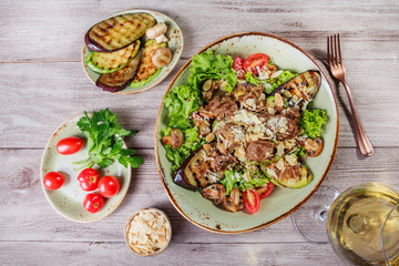 Hot salad with veal, mushrooms, salad leaves, eggplant, zucchini, tomatoes, garnished with grated almonds and Parmesan cheese and glass of wine on wooden background. Healthy food