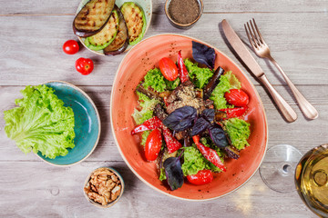 Hot salad with grilled tomatoes, eggplant, zucchini, red pepper, salad leaves, garnished with grated walnuts and basil and glass of wine on wooden background. Italian food. Top view