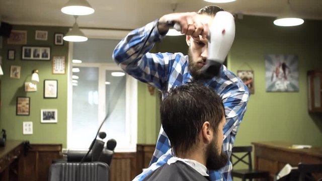 Attractive young barber is cutting human hair with the scissors. He is looking at hair with concentration. The bearded man is raising his chin