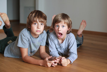 Two surprised kids lying on wooden floor and using mobile phone 
