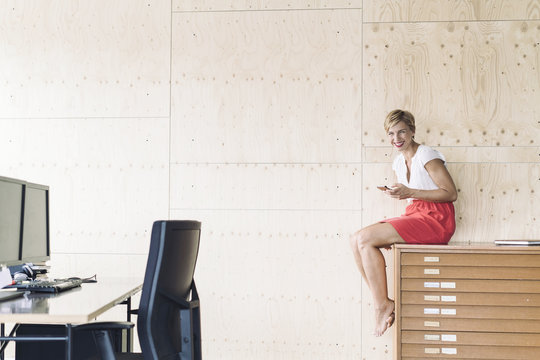 Smiling businesswoman sitting on filing cabinet in creative office