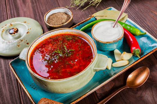 Ukrainian and Russian traditional beetroot soup - borscht in clay pot with sour cream, spice, garlic, pepper, dried herbs and bread on dark wooden background, healthy food. Ingredients on table.