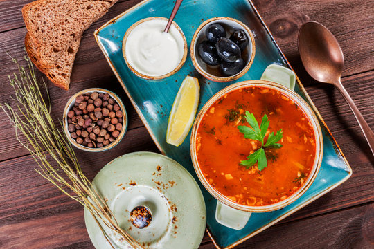 Soup with meat, olives, herbs, lemon, sour cream in bowl, black bread and spices on dark wooden background, homemade food. Traditional Russian soup - solyanka. Top view