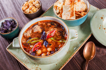 Soup with meat in bowl with oregano, chickpeas, peppers and vegetables served with crackers and bread on dark wooden background, healthy food. Top view