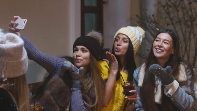 Group of happy female friends drink mulled wine and taking selfie with a smartphone in the outdoor cafe in winter. 60 FPS slow motion, 4K UHD RAW edited footage
