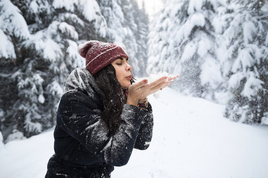 Young Beautiful Woman Blow Snow Hands In Winter Forest Girl Outdoors Walking Snowy White Park Wear Warm Clothes